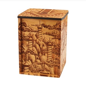 May include: A wooden box with a detailed engraved landscape design featuring towers, mountains, and clouds. The box has a dark border on the lid and is made of light-colored wood.