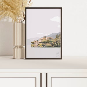 Op de afbeelding: Ingelijste kunstafdruk met een serene landschap met een dorp aan het meer, bergen en een bewolkte lucht. Het kunstwerk is in gedempte tinten grijs, groen en beige. Een hoge, beige vaas met gedroogde planten staat links van de afdruk.