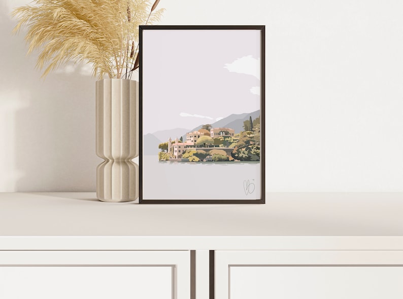 Op de afbeelding: Ingelijste kunstafdruk met een serene landschap met een dorp aan het meer, bergen en een bewolkte lucht. Het kunstwerk is in gedempte tinten grijs, groen en beige. Een hoge, beige vaas met gedroogde planten staat links van de afdruk.