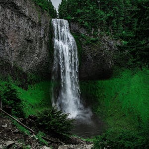May include: A tall waterfall cascades down a steep, dark gray rocky cliff face into a pool below. The surrounding landscape is filled with dense, vibrant green coniferous trees and lush vegetation under a cloudy, overcast sky.
