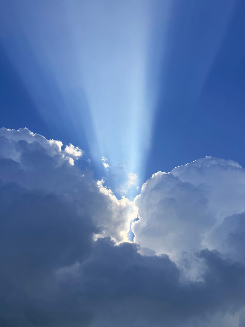 May include: Bright white sunbeams radiate upwards from a gap between two large, dense, dark blue and white cumulus clouds against a clear, deep blue sky. The light creates a dramatic contrast, highlighting the edges of the cloud formations.