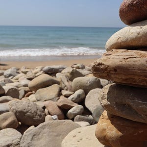 May include: A vertical stack of smooth, rounded, earth-toned stones in shades of tan, beige, and brown. The stones are layered on top of each other, creating a textured column that stands in the foreground against a blurred beach landscape.