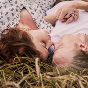 May include: A white short-sleeved shirt with a repeating black cat pattern and a plain white short-sleeved shirt worn by two individuals lying down in dry grass, with one person wearing a ring on their finger and holding the other's hand.