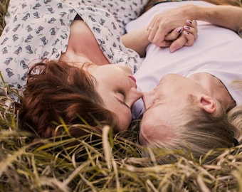 May include: A white short-sleeved shirt with a repeating black cat pattern and a plain white short-sleeved shirt worn by two individuals lying down in dry grass, with one person wearing a ring on their finger and holding the other's hand.