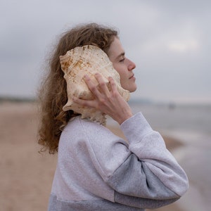 May include: A person with curly brown hair holds a large seashell to their ear on a sandy beach. The sky is overcast and the ocean is in the background.