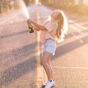 May include: A woman in a light pink shirt and denim shorts pops a bottle of champagne on a paved road. The champagne spray is visible in the air.