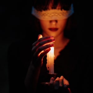 May include: A person with a white blindfold holds a lit candle in a dark setting.