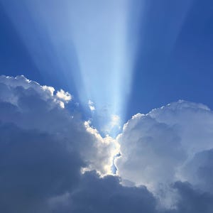 May include: Bright white sunbeams radiate upward from a gap between two large, dense, dark blue and white cumulus clouds against a clear, deep blue sky. The light creates a dramatic contrast, highlighting the edges of the cloud formations.