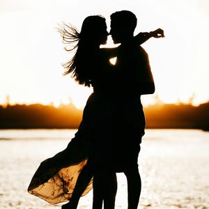 May include: A silhouette of two people embracing against a bright sunset. One person has long hair blowing in the wind and wears a flowing dress with a floral pattern, while the other person stands close, with their arms wrapped around the first person.