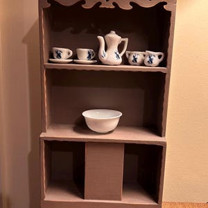 Può includere: Un piccolo mobile in legno dipinto di marrone chiaro, con ripiani e un vano inferiore. I ripiani espongono un servizio da tè bianco con disegni floreali blu, tra cui una teiera, tazze e piattini, insieme a una piccola ciotola bianca.