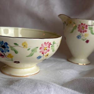 May include: A vintage cream-colored sugar bowl and creamer set with a floral design. The set features delicate pink, blue, and yellow flowers with green leaves on a cream background.