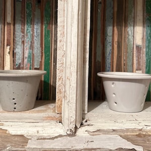 Op de afbeelding: Twee kleine, lichtgrijze keramische potten met een uitlopende vorm en een reeks kleine gaatjes. De potten worden getoond in een rustieke houten kast met een verweerde witte lijst en een kleurrijke, verweerde achtergrond.