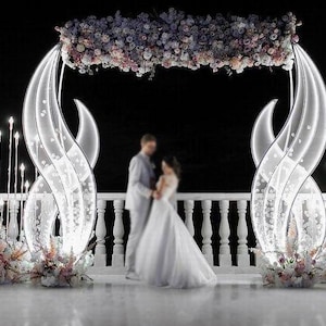 以下が含まれることがあります： エレガントな結婚式のセレモニーセットアップ。花柄のアーチと装飾的な照明が特徴です。カップル、白い手すり、照らされた抽象的な構造物があります。ピンクと白の花の装飾がアクセントになっています。