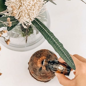 Op de afbeelding: Een bruine kokosnootschelpdiffuser met een klein bruin glazen flesje etherische olie dat in de bovenkant wordt gegoten. De diffuser wordt omringd door een boeket witte en groene bloemen.