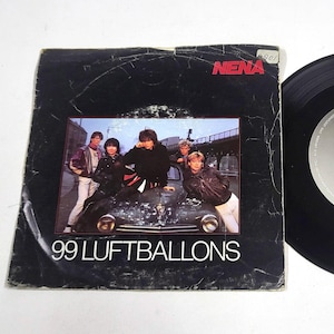 May include: Vintage vinyl record and sleeve for the song "99 Luftballons" by Nena. The black sleeve features a photo of the band and the band's name in red. The record is black with a silver label.
