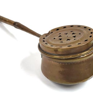 Puede incluir: Una olla antigua de color cobre con un mango de madera. La olla tiene una tapa redonda perforada y un cuerpo curvo. El mango está unido al costado y se extiende hacia afuera. Probablemente utilizado para cocinar o calentar.