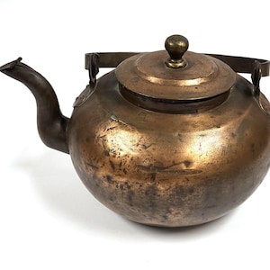 Puede incluir: Una tetera de cobre vintage con un acabado desgastado y una tapa. La tetera tiene un pico curvo y un asa con un pomo decorativo.
