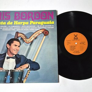 May include: Vintage vinyl record album cover featuring Luis Bordon, a Paraguayan harp soloist. The album cover has text in pink and yellow, with a photo of Bordon playing the harp and drums. A black vinyl record is next to the album cover.