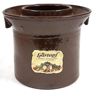 Puede incluir: Un tarro de fermentación de cerámica marrón con un borde ancho y una depresión central. El tarro tiene una capacidad de 3,5 litros y presenta una etiqueta con el texto "Gärtopf" y una ilustración de verduras. Tiene dos asas laterales.