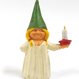 May include: A whimsical figurine of a woman wearing a green pointed hat and a white dress. She holds a white candle in a red holder. The figurine has blonde hair and a smiling expression.