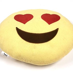 Peut inclure: Un coussin emoji jaune avec un visage souriant et des yeux en forme de cœur rouges. Le coussin est rond et a une surface douce et texturée. La bouche est de couleur marron foncé.