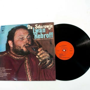 Può includere: Una copertina di un album in vinile vintage con un uomo che beve da un bicchiere. La copertina dell'album presenta un testo in tedesco, con il nome dell'artista "Iwan Rebroff" e il titolo "Da Sdarowje". Il disco è nero con un'etichetta arancione.