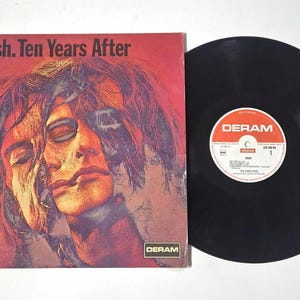 Puede incluir: Un álbum de vinilo vintage de Ten Years After, titulado "Ssssh.", con una portada roja y naranja que presenta un retrato surrealista. El disco negro está junto a la portada del álbum. La etiqueta del disco es visible.