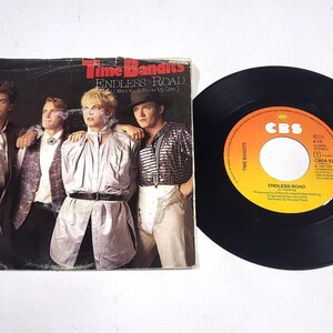 Könnte beinhalten: Eine Vintage-Vinyl-Schallplatte und das Albumcover für den Song "Endless Road" von Time Bandits. Das Albumcover zeigt eine Gruppe von vier Personen in Kleidung der 1980er Jahre. Das Plattenlabel ist orange und schwarz.