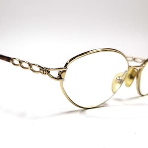 May include: Gold-toned eyeglasses with oval lenses. The frames feature a chain-link design on the temples and a subtle CD logo. The earpieces are a darker brown color. The glasses are set against a white background.