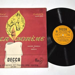 以下が含まれることがあります： プッチーニのオペラ「ラ・ボエーム」のヴィンテージビニールレコード。アルバムの表紙は赤色で、ろうそくの形をしたイラストと「Puccini's La Bohème」の文字が描かれています。レコードのラベルは黄色で黒い文字が書かれています。レコードは黒色です。