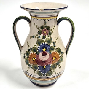 May include: A cream-coloured ceramic vase with two handles and a wide opening. The vase is decorated with a floral design featuring blue, orange, pink, and purple flowers with green leaves. The handles are green with blue trim.
