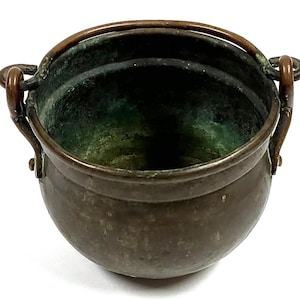 Pode incluir: Um pequeno caldeirão antigo de bronze com corpo arredondado e abertura larga. Possui duas alças curvas nas laterais e uma alça resistente na parte superior. O interior mostra uma pátina verde e o exterior tem uma aparência envelhecida.