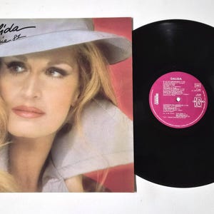 May include: Album cover featuring Dalida, with the text "Palida Olympia 81" in script. The album cover has a portrait of Dalida wearing a hat. A black vinyl record is next to the album cover.
