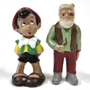 May include: Two vintage figurines: Pinocchio in a red hat, yellow shirt, green vest, and black shorts, and an older man with a white beard, green shirt, brown vest, and red pants. Both figures are painted with bright colors.