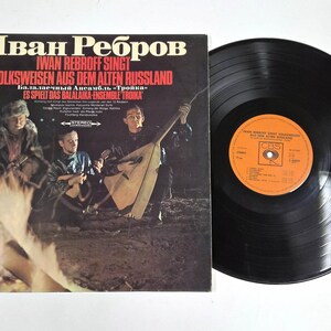May include: An album cover and vinyl record. The album cover features a group of musicians playing instruments around a campfire. The album title is in Russian and German. The vinyl record is black with an orange label.
