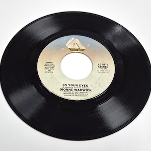 Puede incluir: Un disco de vinilo negro con una etiqueta blanca y beige. La etiqueta presenta el texto "IN YOUR EYES" de Dionne Warwick, producido por Barry Manilow. El logotipo de Arista Records es visible.