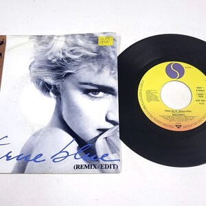 Peut inclure: Un disque vinyle et une pochette d'album pour "True Blue (Remix/Edit)" de Madonna. La pochette de l'album présente une image en noir et blanc de Madonna. Le disque est noir avec une étiquette jaune et violette.