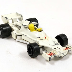 Puede incluir: Un coche de carreras de juguete blanco vintage con neumáticos negros y un piloto amarillo. El coche presenta un alerón trasero y banderas de la Union Jack en la parte delantera. Un juguete coleccionable.