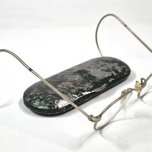 Peut inclure: Lunettes en métal argenté avec des montures fines et des branches incurvées, placées devant un étui rigide ovale présentant une finition texturée marbrée vert foncé et noir avec une bordure argentée métallique le long de la fermeture centrale.