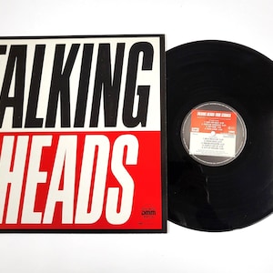 Peut inclure: Un disque vinyle noir avec une pochette d'album rouge et blanche sur laquelle est inscrit "Talking Heads" en lettres noires grasses. L'étiquette du disque est visible et indique "Talking Heads -  More Songs About Buildings and Food".