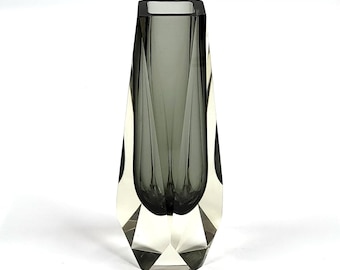 Könnte beinhalten: Eine dunkelgraue Glasvase mit geometrischem Design. Die Vase hat eine quadratische Öffnung und eine spitze Basis.