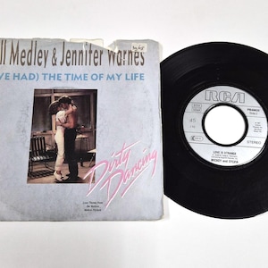 Puede incluir: Un disco de vinilo de 45 RPM vintage y su funda para la canción "(I've Had) The Time of My Life" de Bill Medley & Jennifer Warnes. La funda presenta una foto de una pareja bailando y el título de la película "Dirty Dancing".
