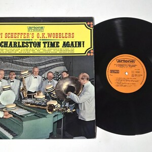 May include: Vintage vinyl record album cover for "It's Charleston Time Again!" by Pi Scheffer's O.K. Wobblers. The cover features a band in suits and bow ties, playing instruments. The record itself is black with an orange label.