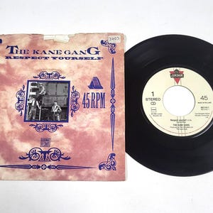 Peut inclure: Un disque vinyle vintage et sa pochette pour "The Kane Gang - Respect Yourself". La pochette rose présente des éléments décoratifs bleus et une photo. Le disque noir est à côté de la pochette.