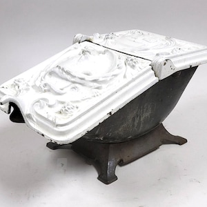 May include: Antique cast iron coal scuttle with a hinged lid. The lid is white with ornate relief detailing, and the body is dark gray. The scuttle sits on a square base with four feet. A decorative piece for home decor.
