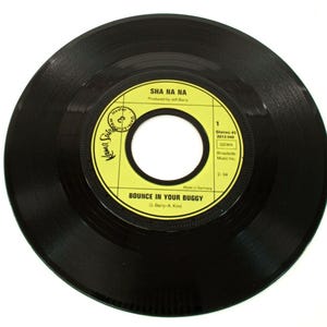 May include: A black vinyl record with a yellow label featuring the text "SHA NA NA" and "BOUNCE IN YOUR BUGGY." The label also includes the text "Made in Germany" and the record's duration, 2:34.