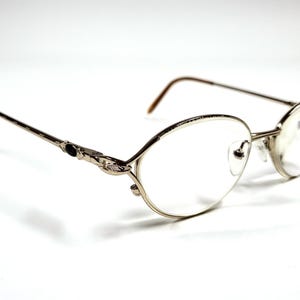 Puede incluir: Gafas plateadas con lentes ovaladas y patillas finas y decorativas. Las monturas tienen un diseño delicado y adornado cerca de las bisagras y puntas de patillas marrones. Las gafas están sobre un fondo blanco.