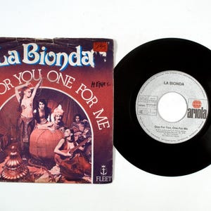 May include: A vintage vinyl record and its sleeve. The sleeve features the text "La Bionda" in blue and white, with "ONE FOR YOU ONE FOR ME" in a red arch. The record is black with a white label.