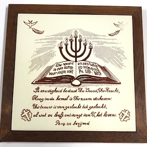 Puede incluir: Una obra de arte religiosa enmarcada con una menorá marrón sobre un libro abierto con texto. La obra de arte incluye texto adicional y elementos decorativos, todo dentro de un marco de madera marrón oscuro. El fondo es blanquecino.