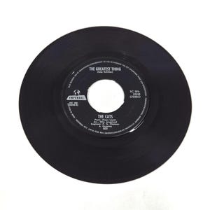 以下が含まれることがあります： 中央に白い文字が記されたラベルを持つ黒い7インチレコード。ラベルにはアーティスト名「The Cats」、曲名「The Greatest Thing」、Imperialのロゴ、そしてKlaas LeyenやWim Jongbloedといった制作クレジットが記載されています。
